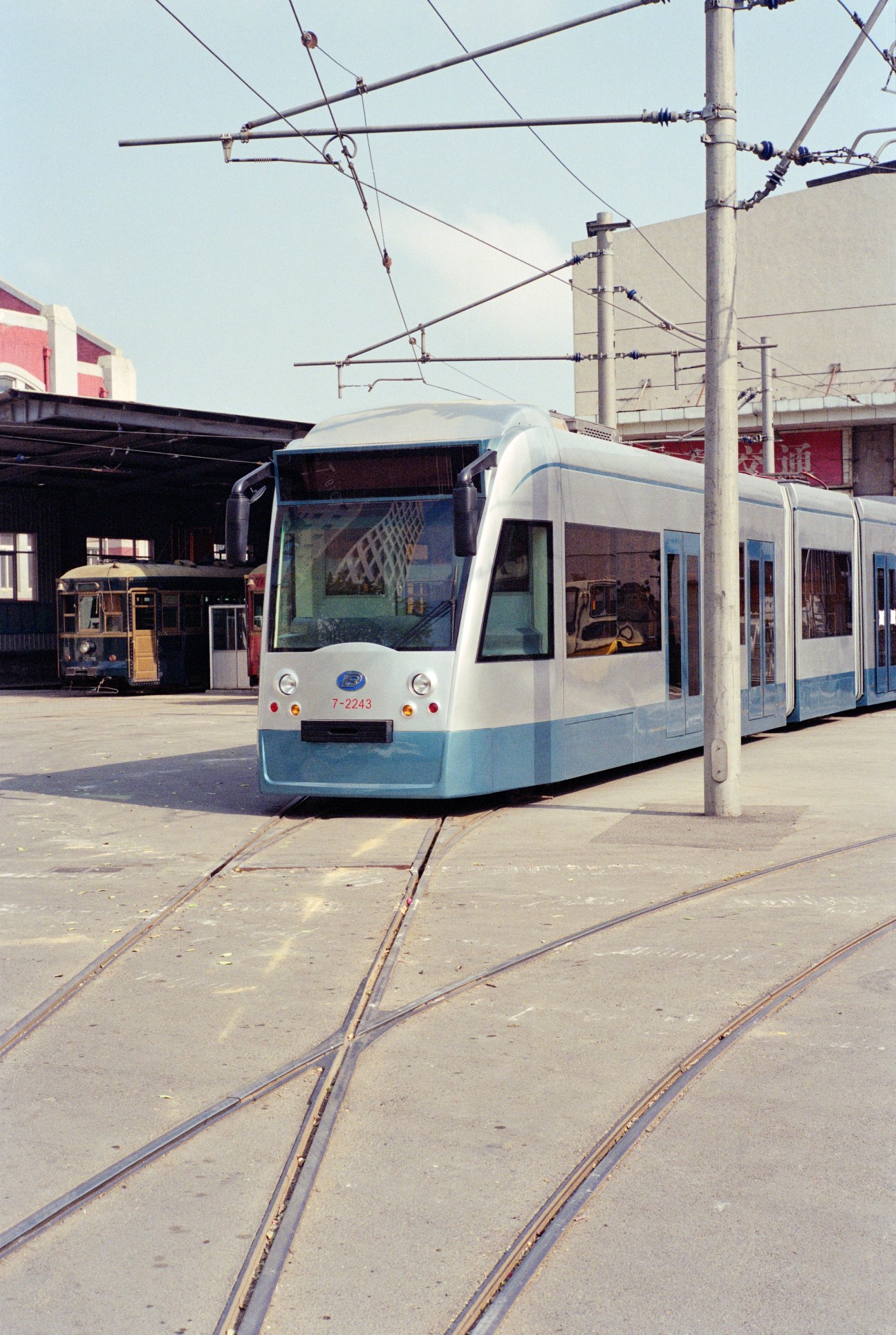 Modern light rail tram on tracks with overhead power lines and vintage trolley visible in background.