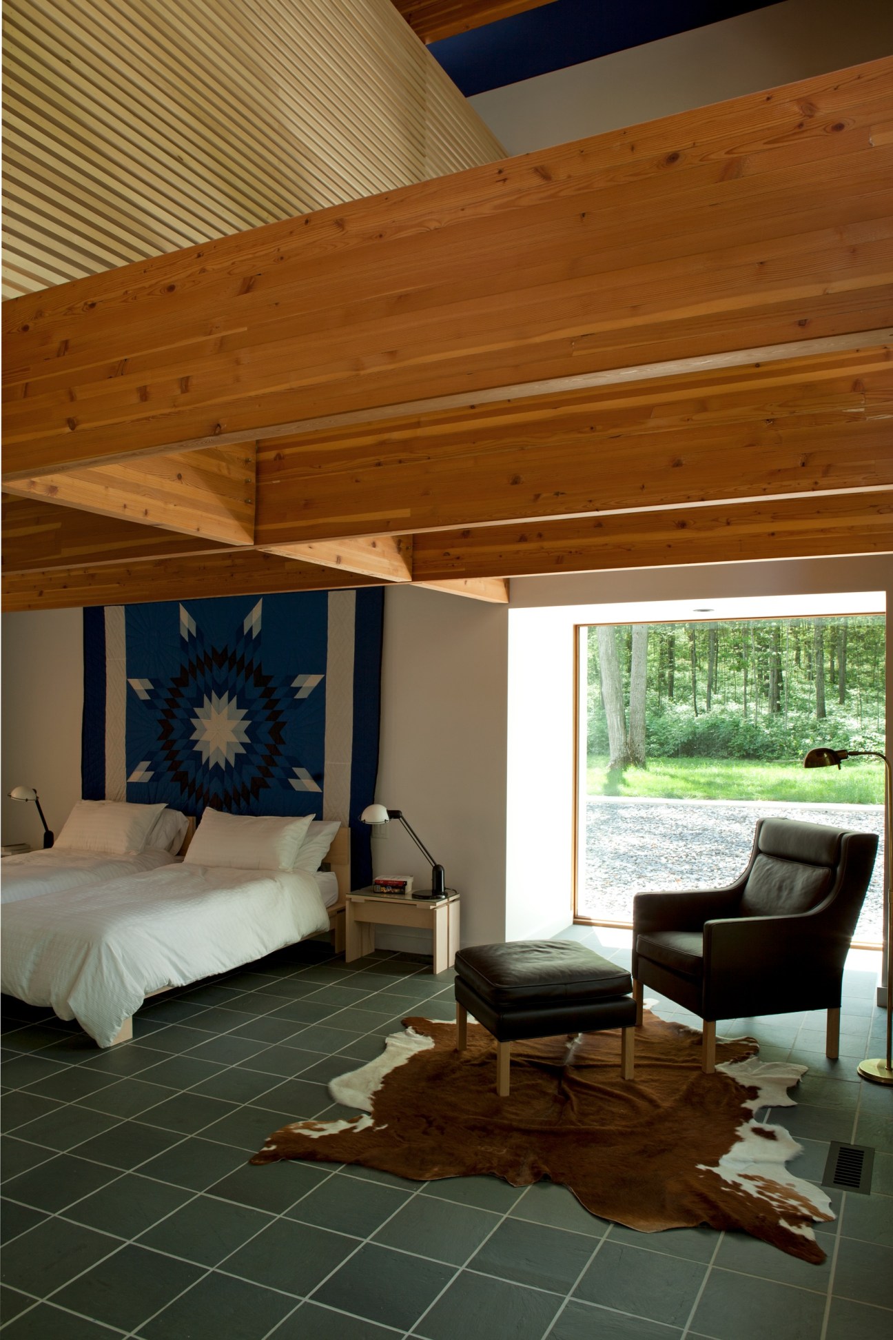 Master bedroom with exposed wood beams, blue geometric wall art, and forest views through large windows.