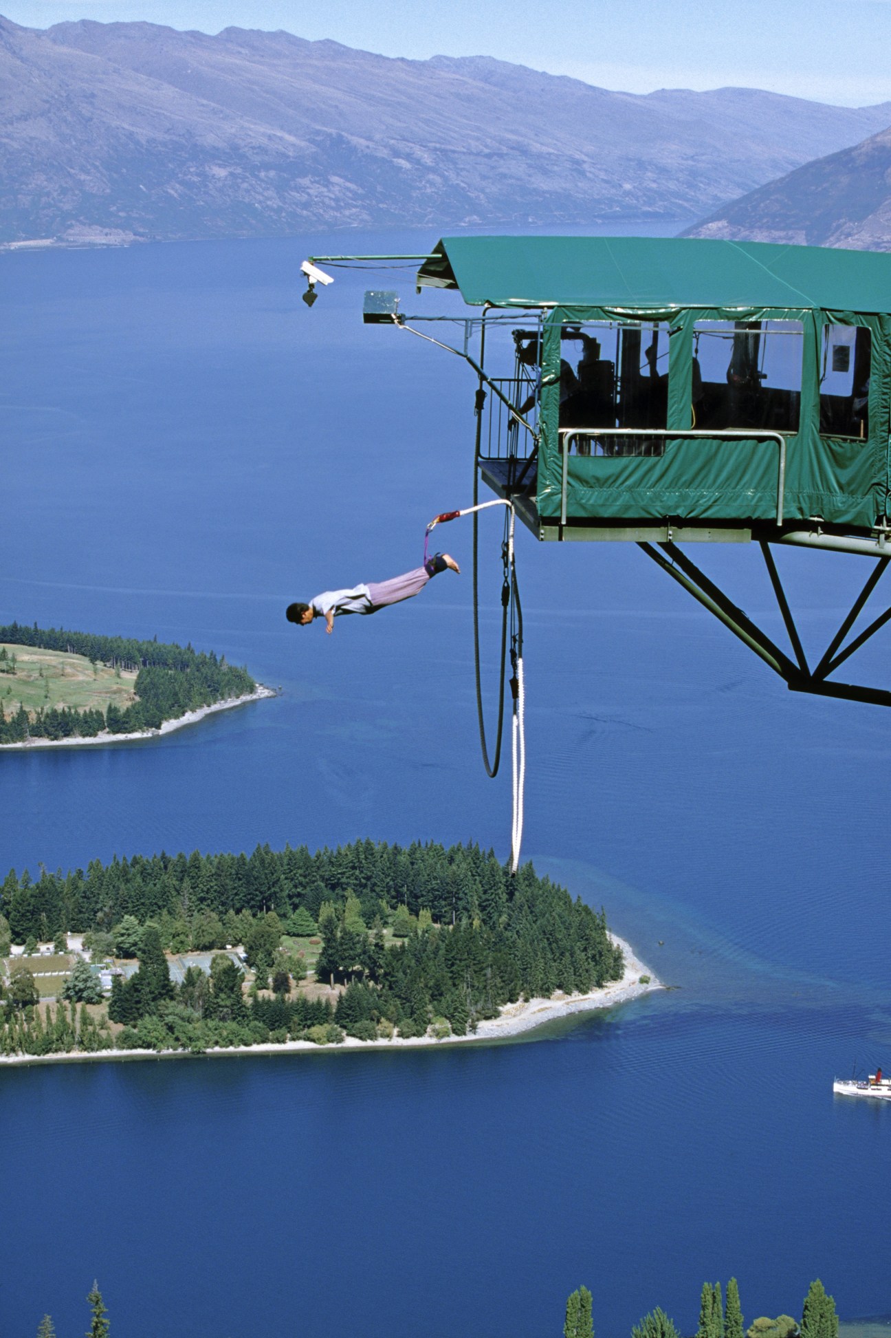 Person bungee jumping from a green cable car platform above a deep blue lake surrounded by mountains and forest.