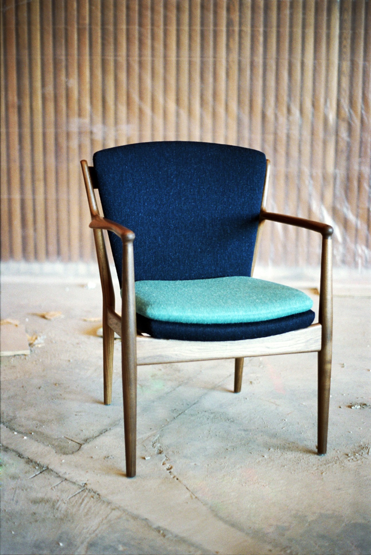 Mid-century modern wooden chair with dark blue backrest and turquoise seat cushion against vertical wood paneling.