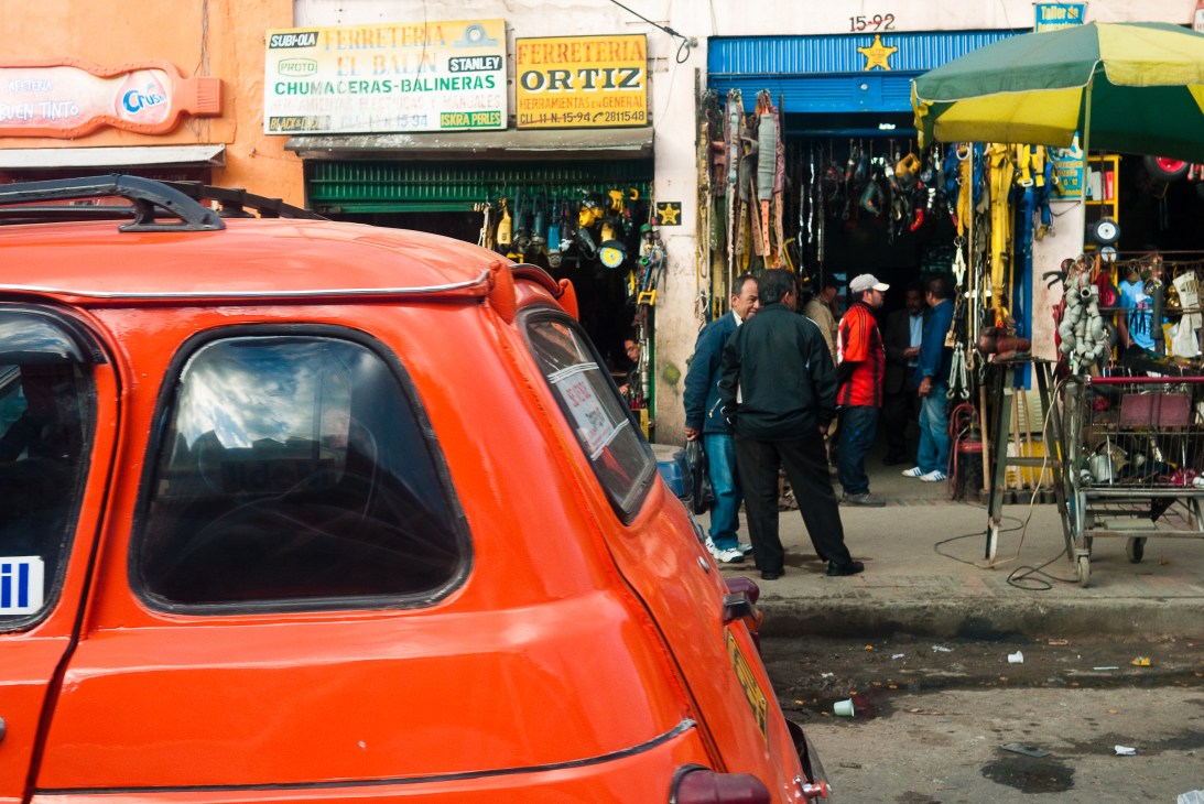 Padre Dario Echeverri conversing with locals on a street in Bogotá's Bronx neighborhood near a red vehicle and shops.