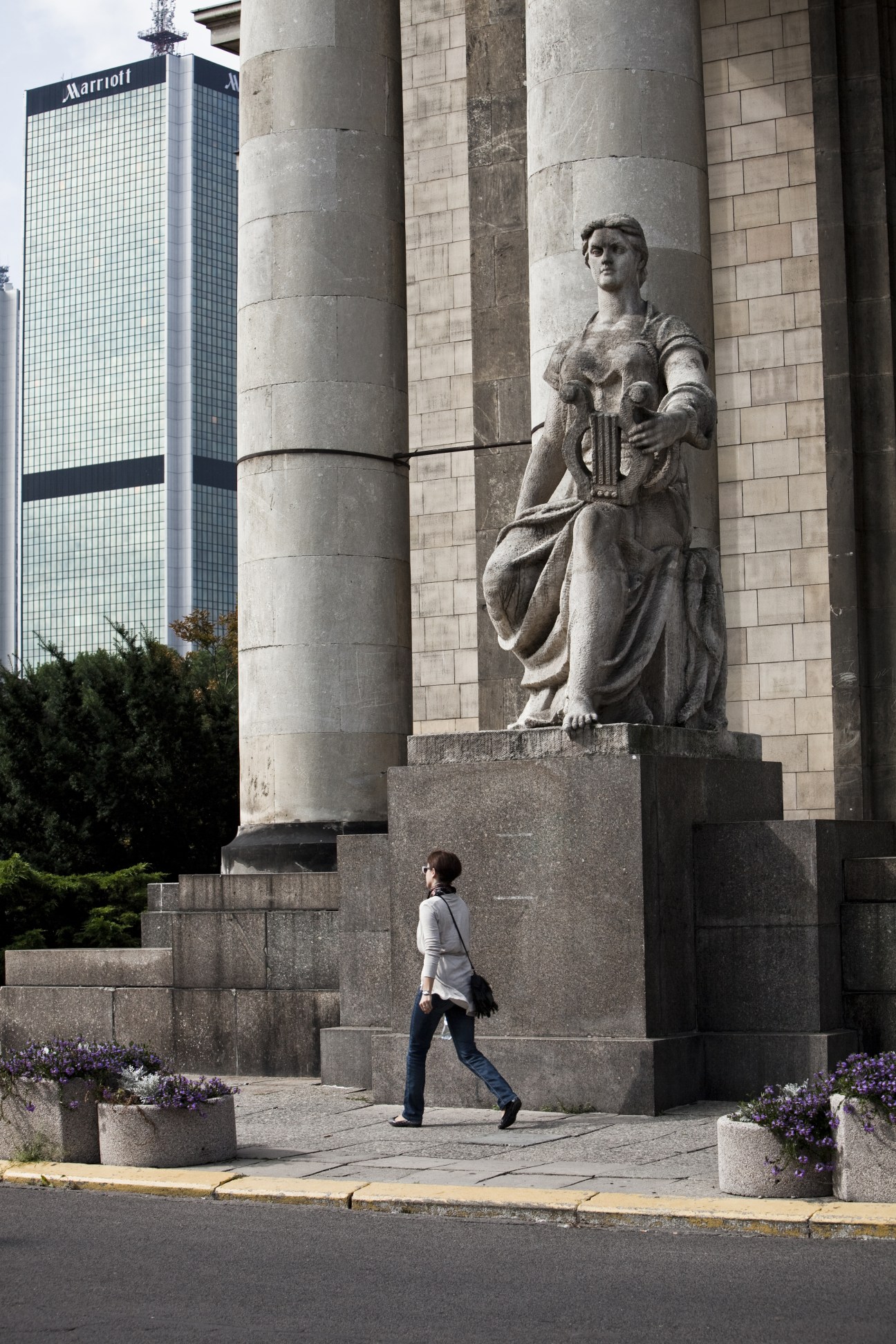 Woman walking past a stone statue at the base of the Palace of Culture and Science in Warsaw, with a modern building visible behind.