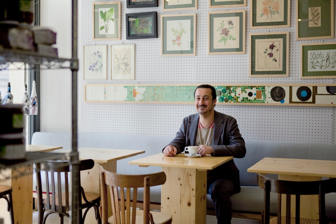 Kamal Mouzawak sits at a wooden table with a cup of coffee in front of a pegboard wall displaying framed botanical prints and decorative items.