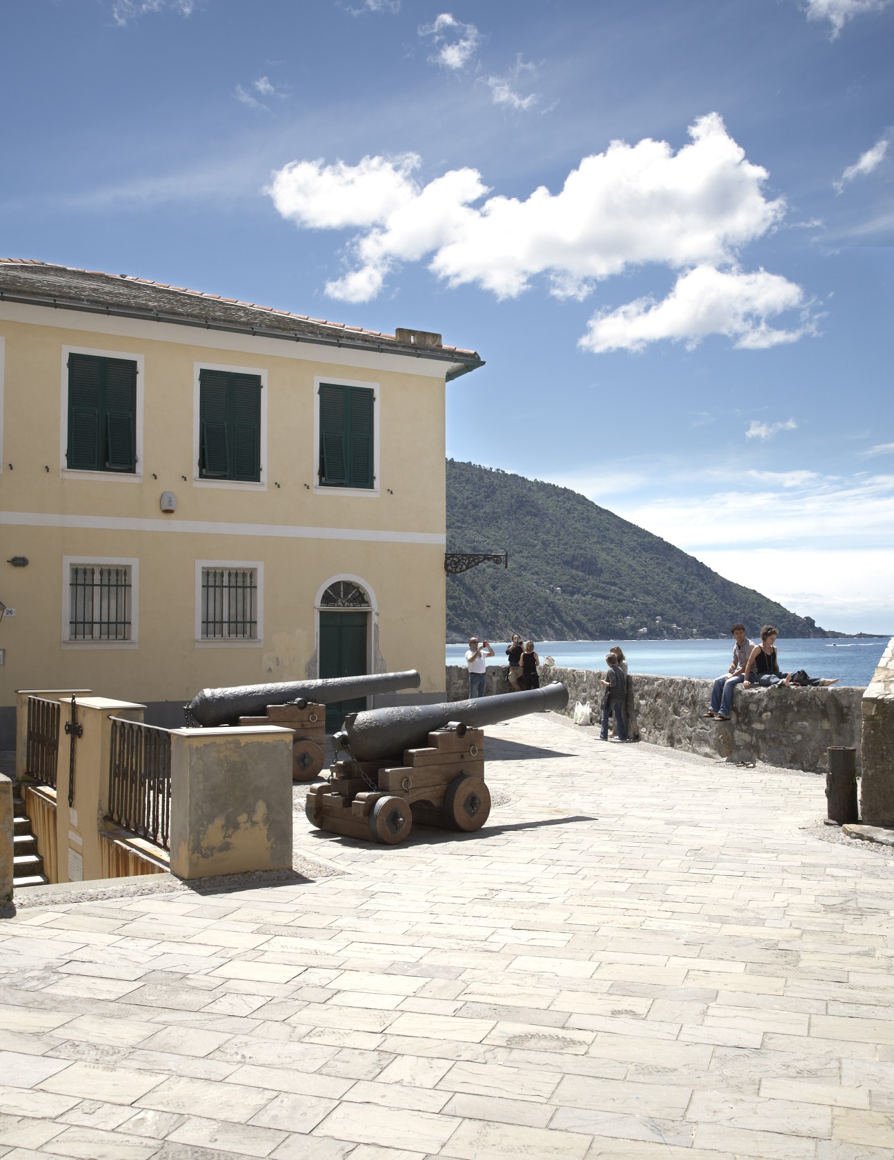 Yellow building with cannons on a waterfront plaza in Camogli, with mountains across the bay.