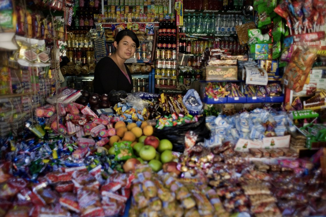A woman stands behind the counter of a colorful street kiosk stocked with candies, snacks, beverages and fresh fruit.