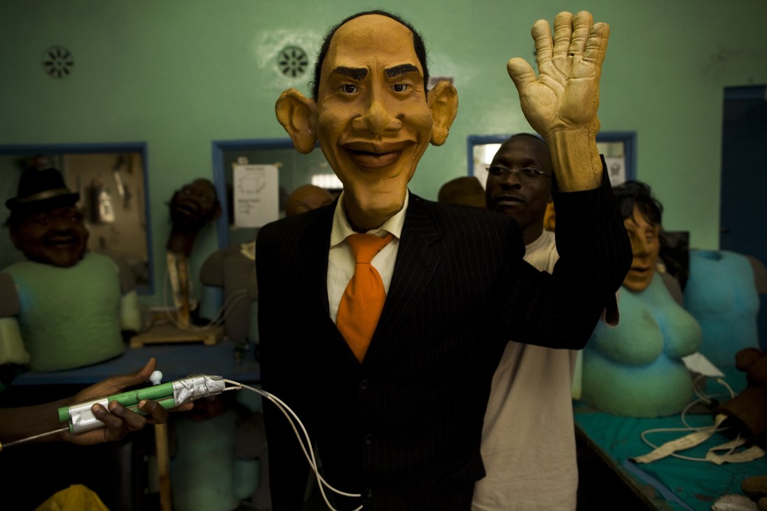 Obama puppet in black suit and orange tie waving, surrounded by film crew in studio