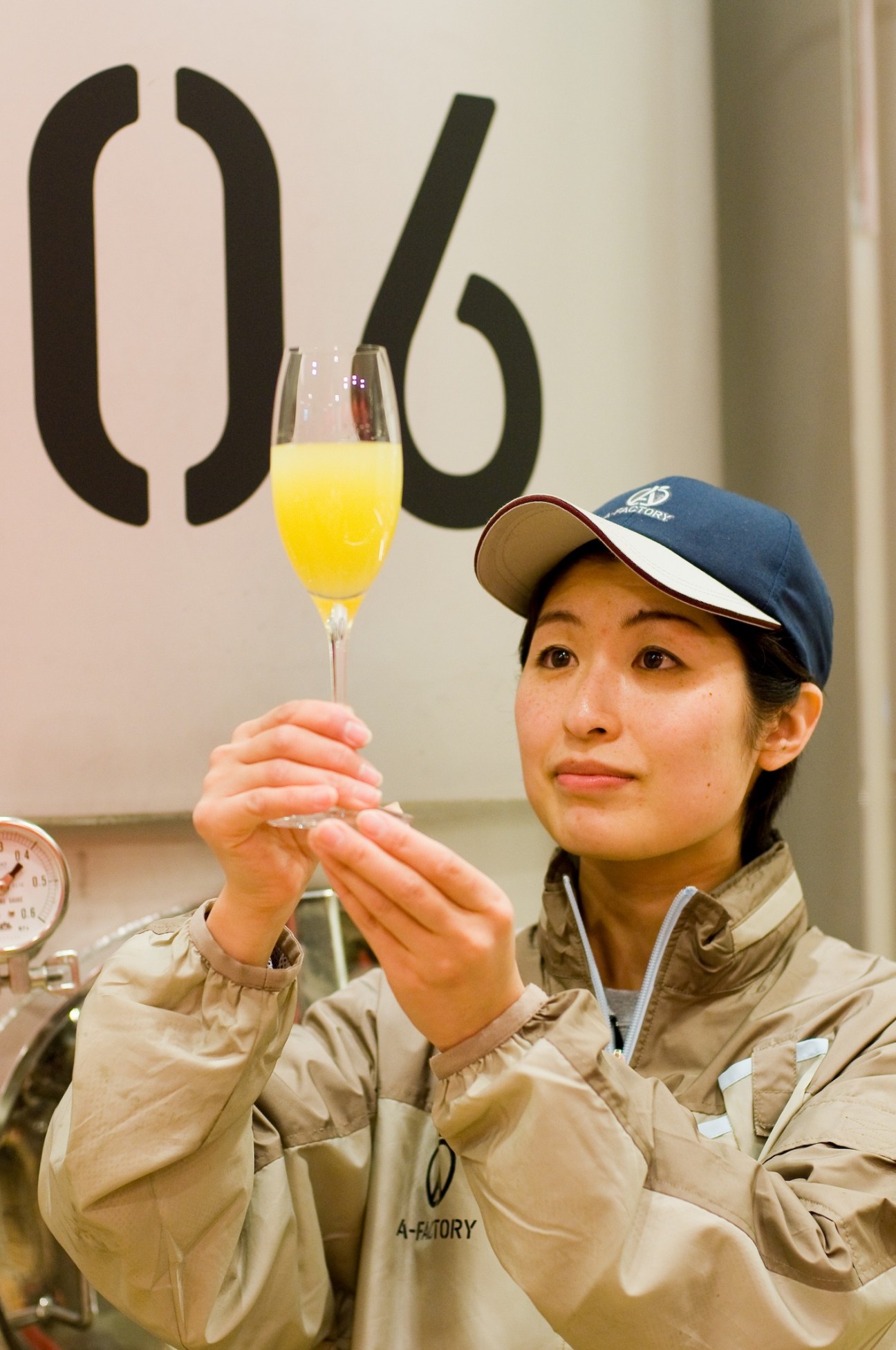 A-Factory worker in tan jacket and blue cap holds a glass of pale yellow cider against a wall with large black numerals.