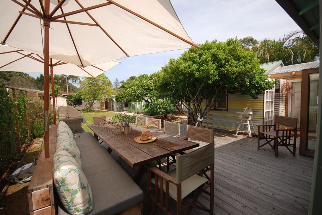 Outdoor dining area with wooden table, chairs, umbrella and fruit trees in the background.