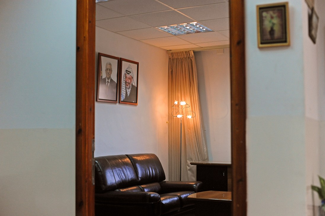 Waiting room with black leather seating, framed portraits on wall, and floor lamp in Palestinian Negotiations Affairs Department office.