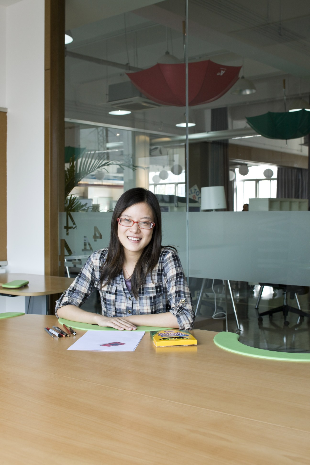 Student Xing Jing Qi, 21, sits at a wooden table in a modern design studio with colorful umbrellas hanging overhead.