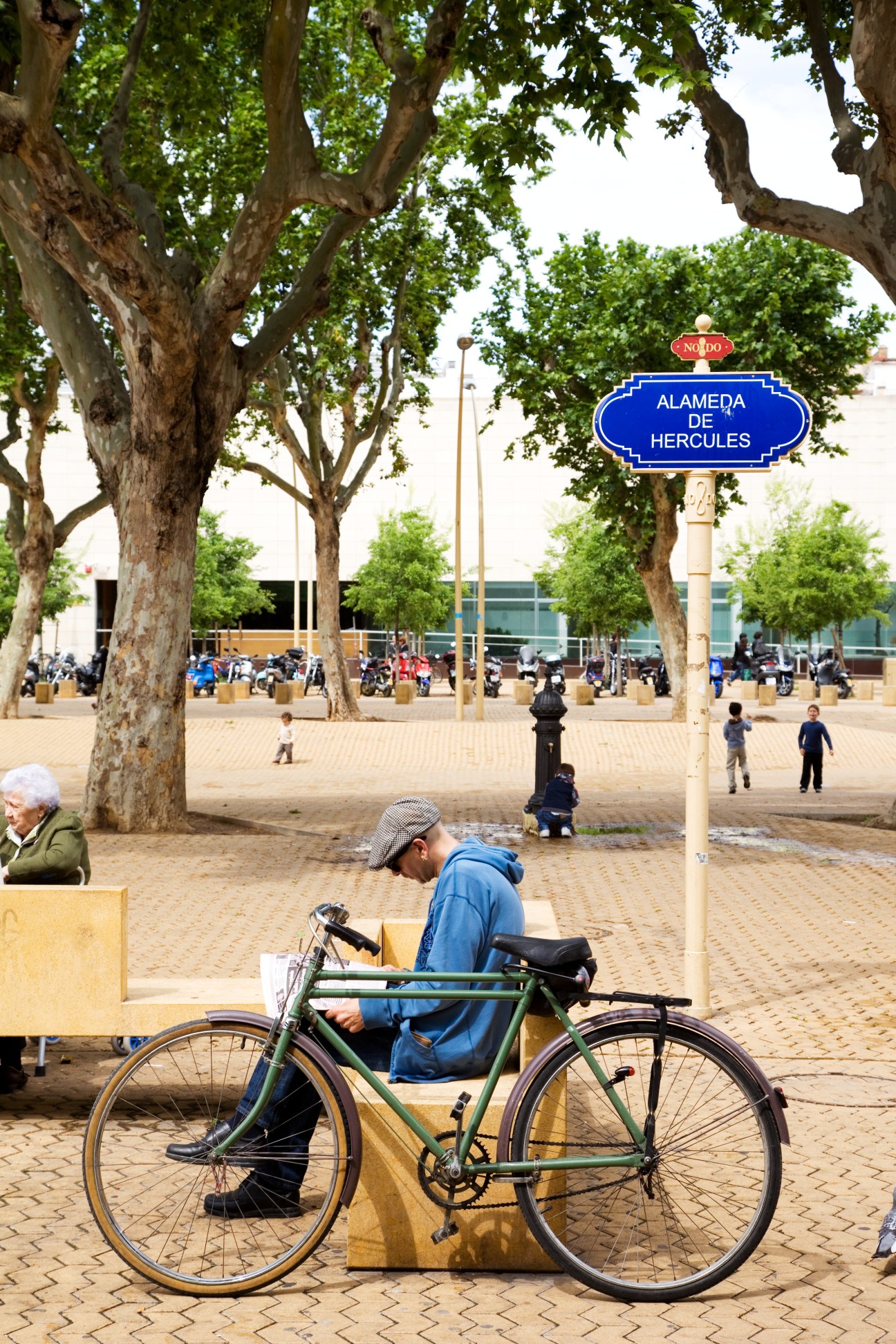 Man sitting on bench with bicycle at Alameda de Hércules park in Seville, with large trees and blue street sign visible.