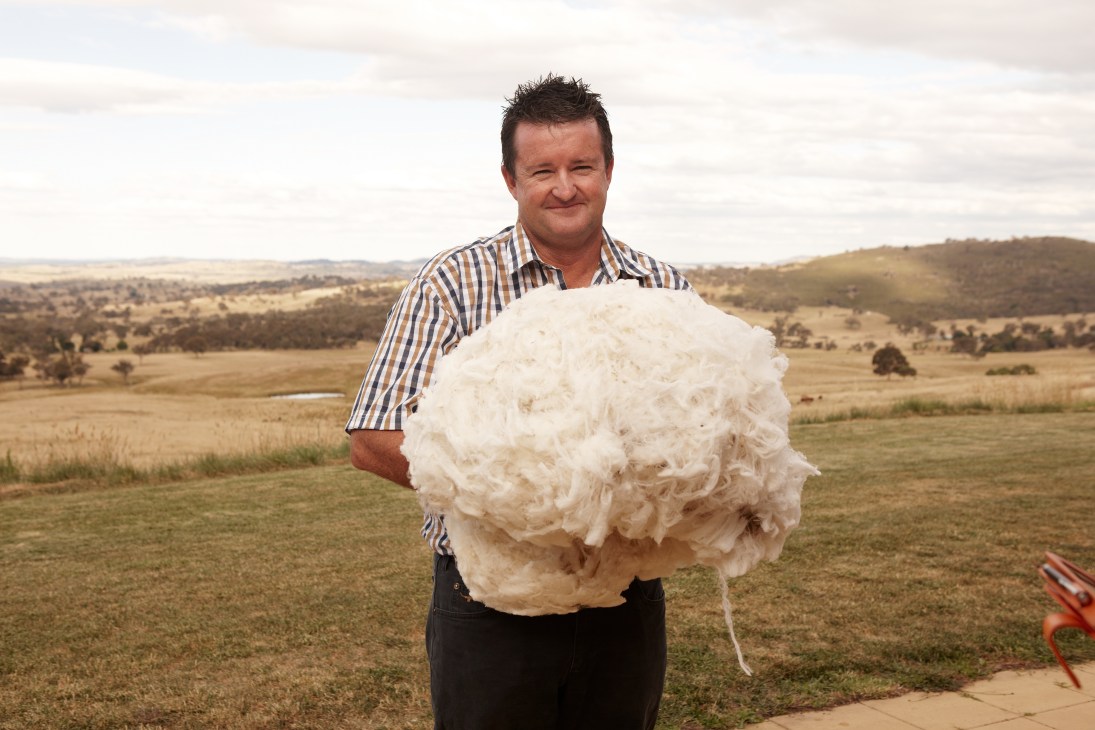 Woolgrower Andrew Ledger holds a large fluffy white wool fleece in a pastoral landscape.