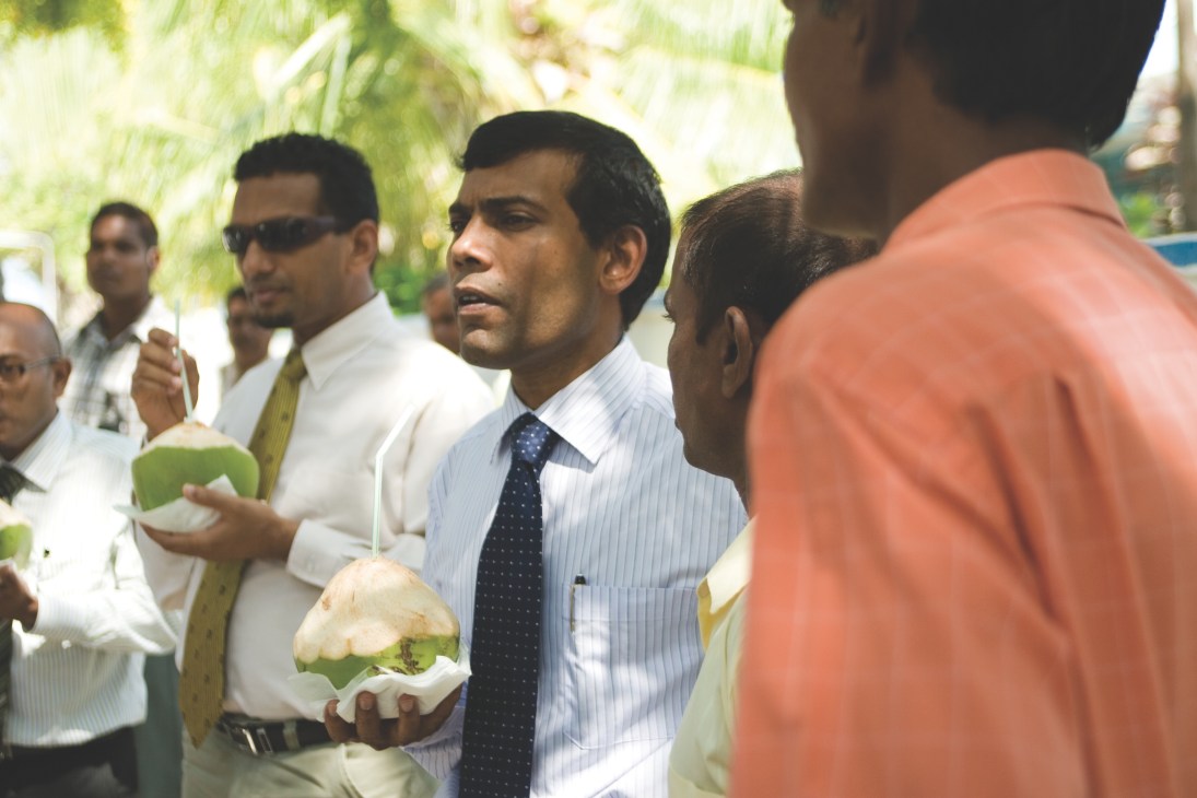 Men in formal attire holding coconuts while conversing outdoors in a tropical setting.