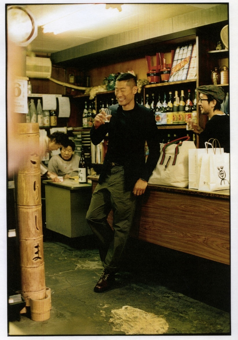 Man in black Dior Homme jacket and Yaeca clothing smiles in a Japanese liquor store interior.