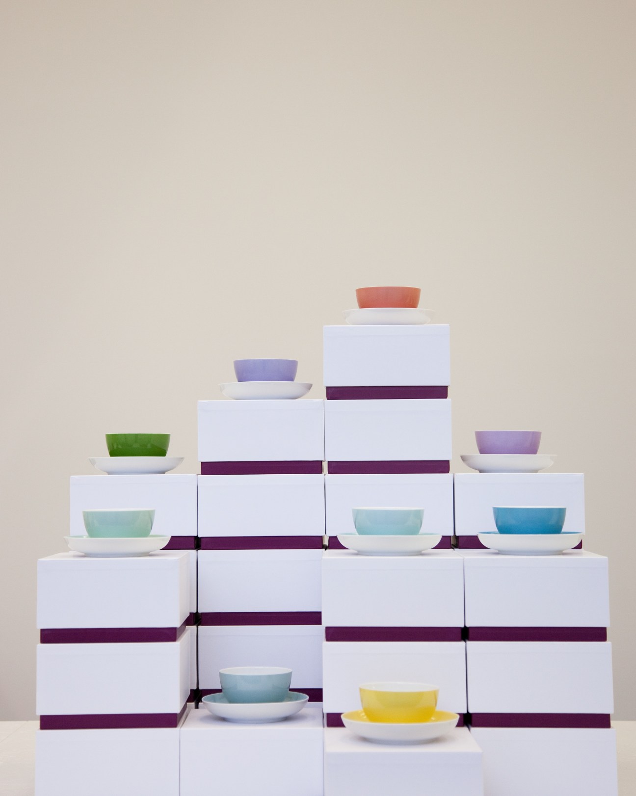 Stacked white boxes with burgundy straps displaying colorful ceramic bowls and saucers in a pyramid arrangement.