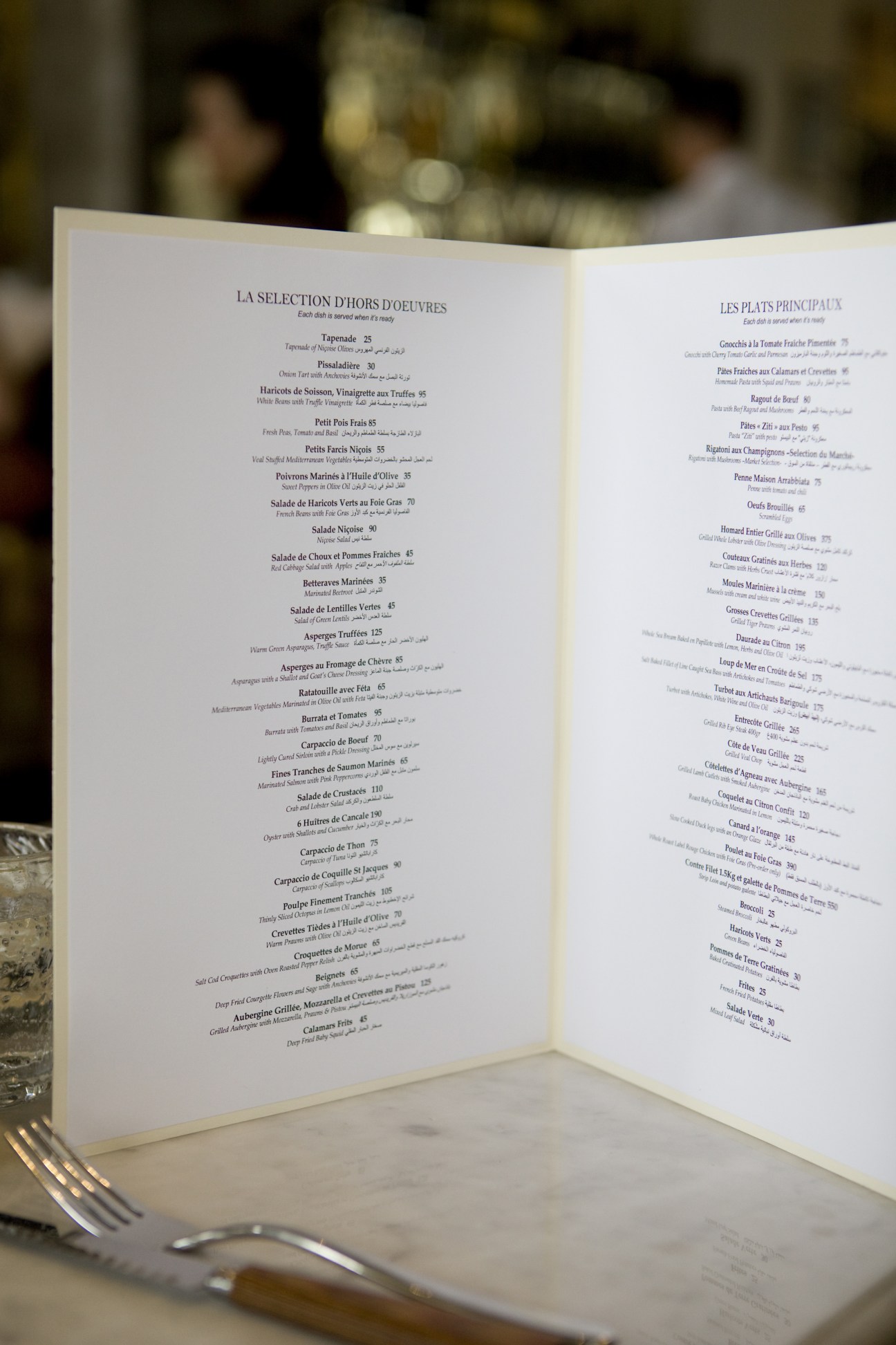 An open French restaurant menu at La Petite Maison with bilingual French and Arabic text listing appetizers and main courses.