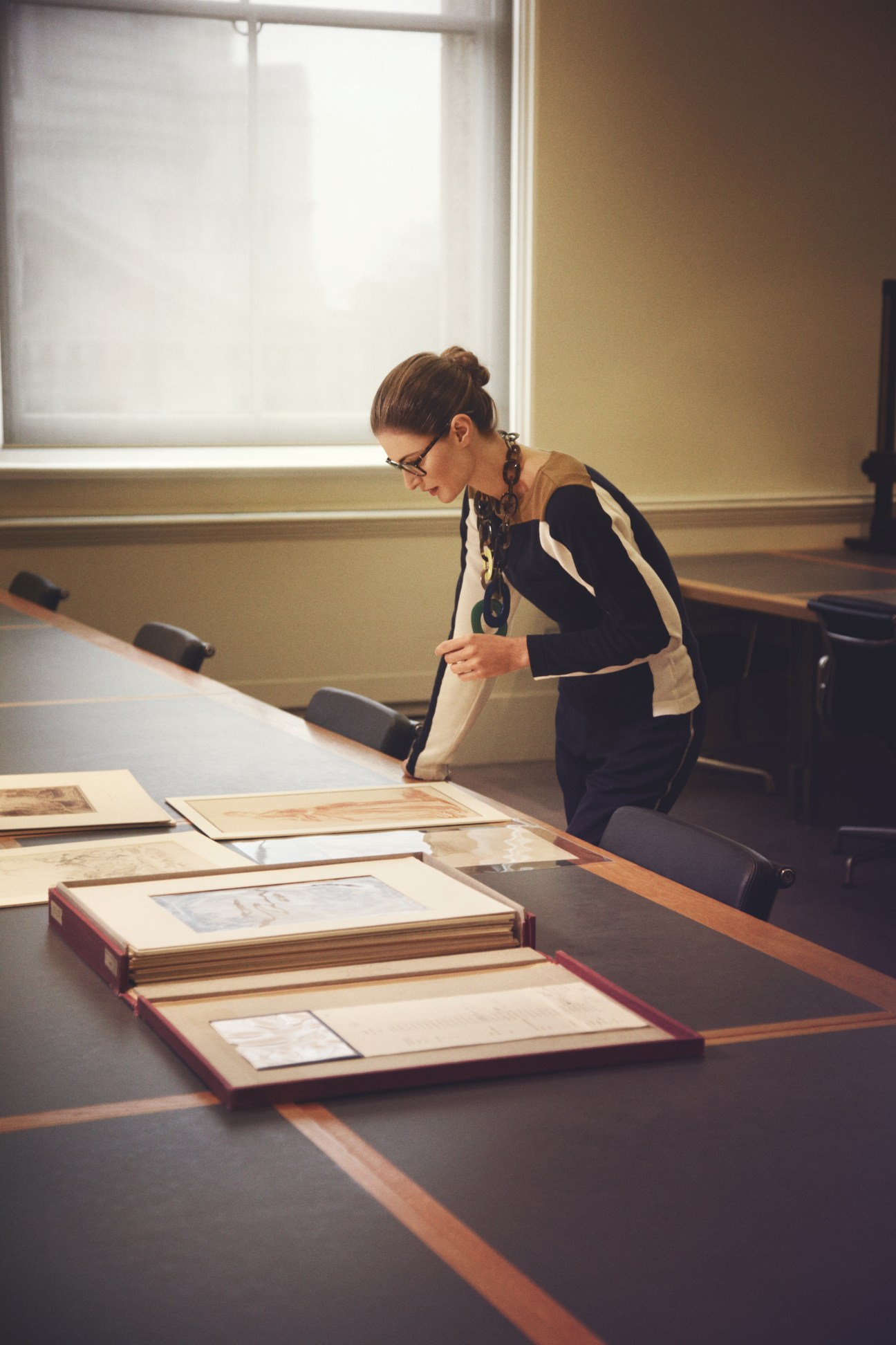 Woman wearing Prada glasses, Hermès necklace, Lacoste jumper and 1205 trousers examining archival documents at a library table.