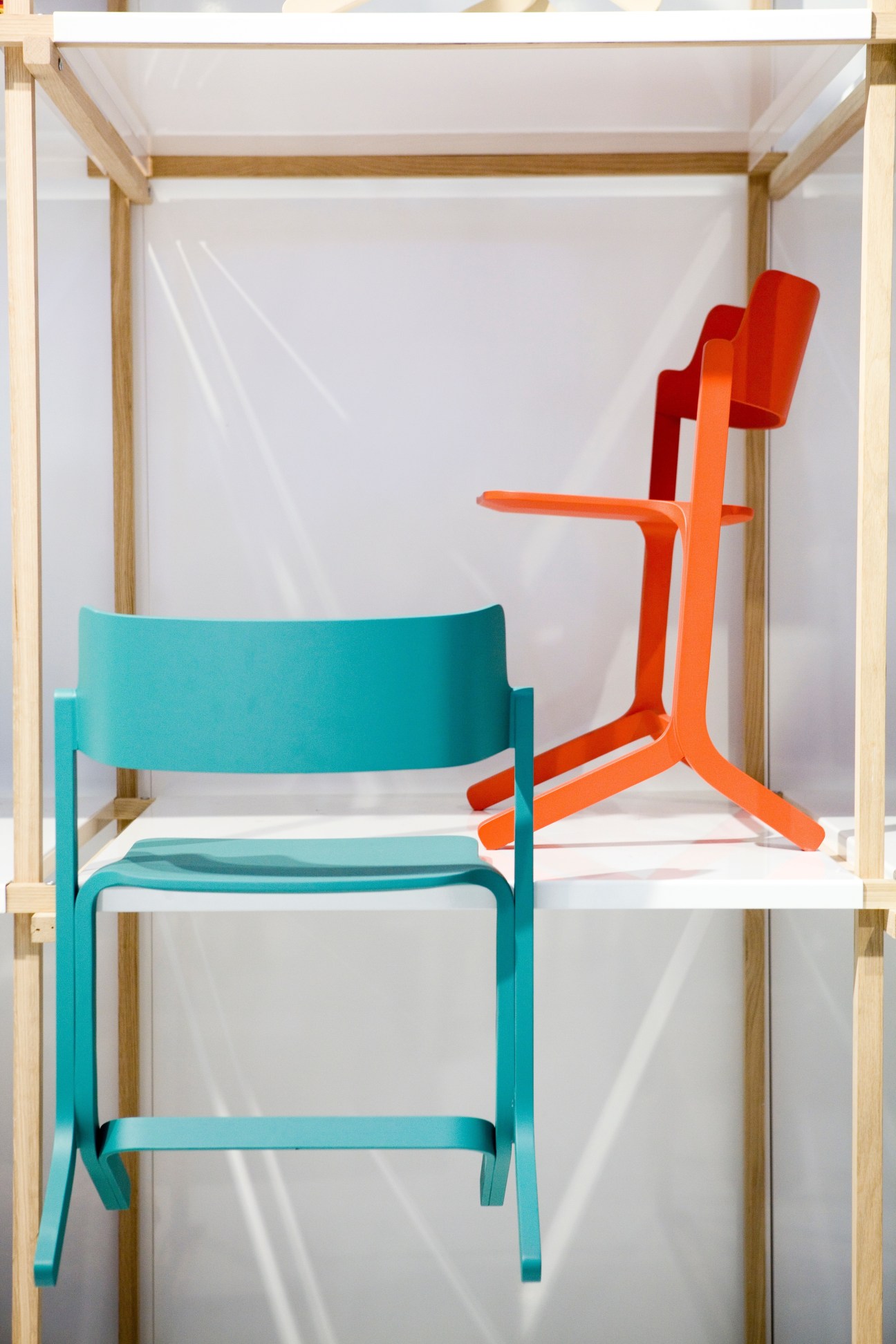 A turquoise curved chair and red sculptural chair displayed in a wooden frame display case.