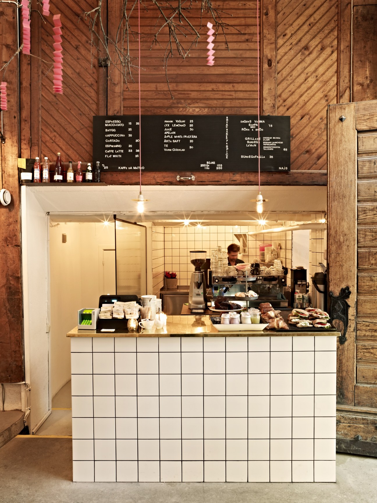 Modern white-tiled café counter with barista working inside, wooden ceiling and rustic brick walls above.