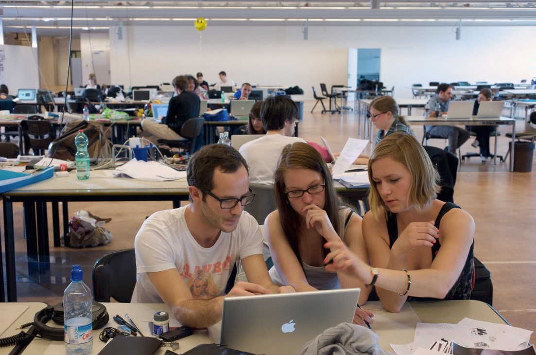 Three design students collaborate at a desk with a laptop in a bright, open studio workspace filled with other students and workstations.