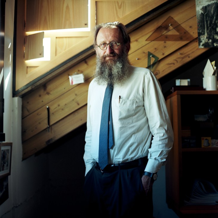 Michele De Lucchi stands in his Milan office with wooden architectural models and natural wood panelling visible behind him.