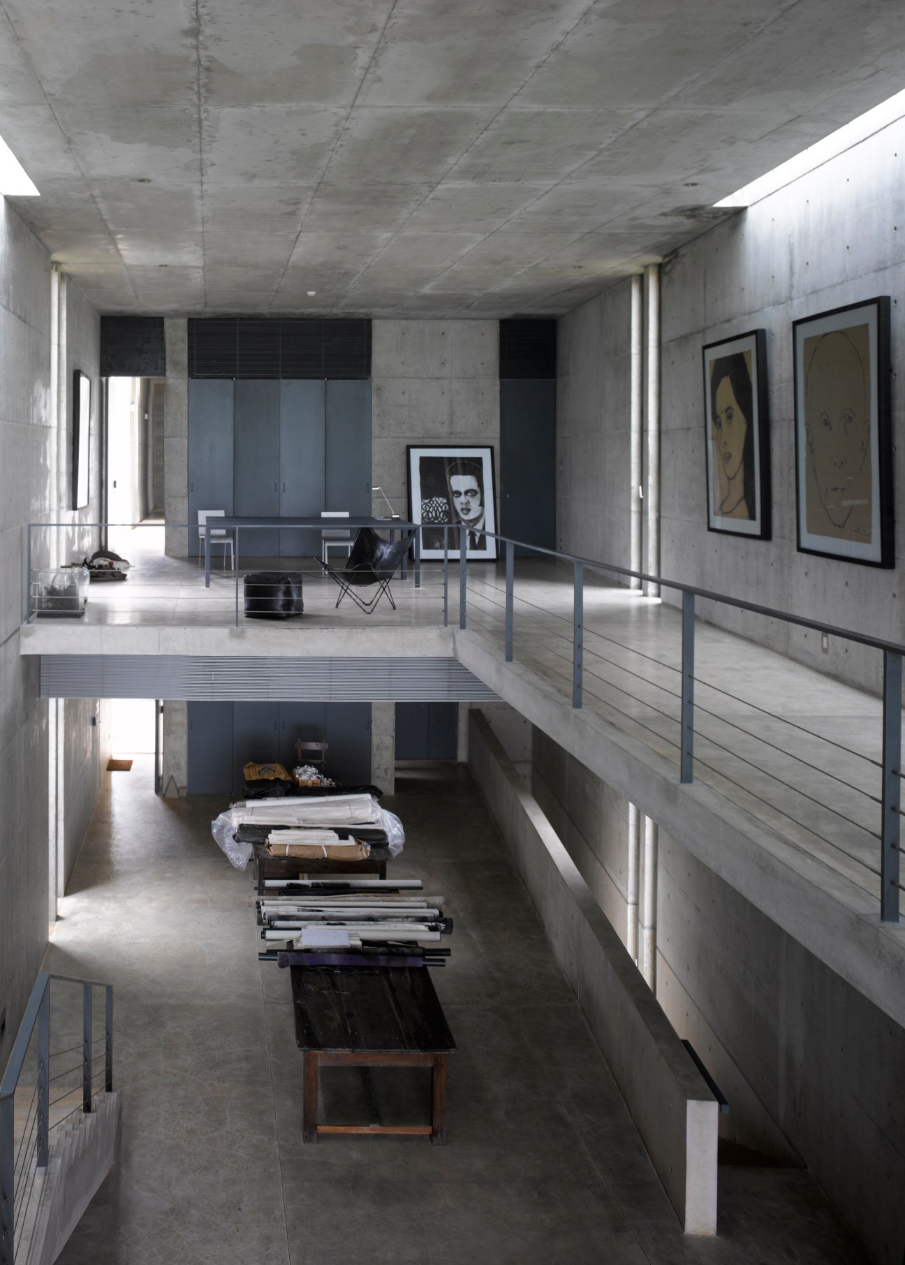 Mezzanine level with railings overlooking artist's workspace below, featuring bare concrete walls, framed artworks, and stacked materials.