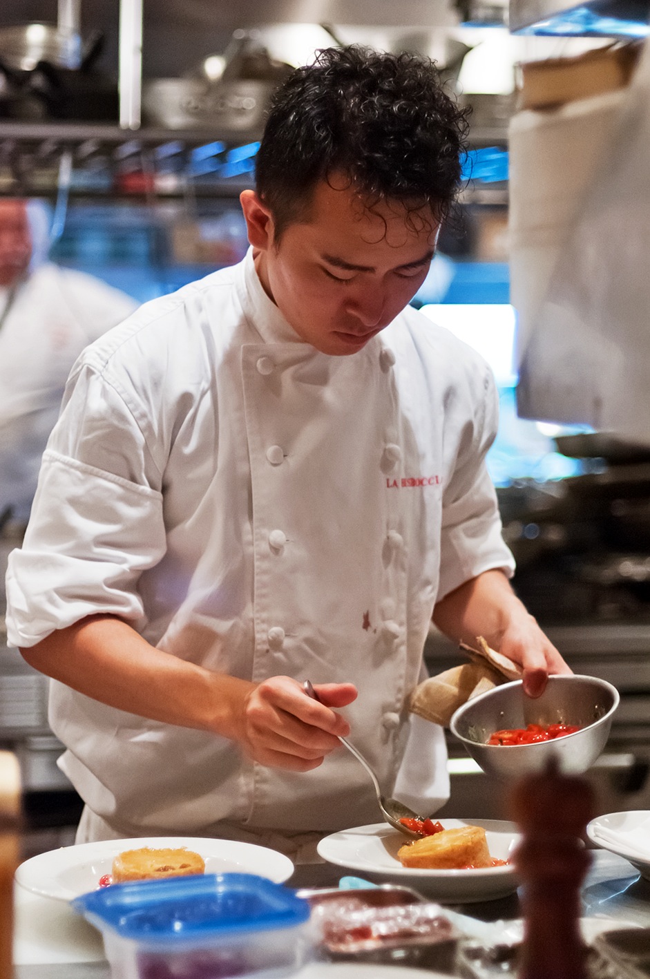 A cook in white chef's jacket plates food in a professional kitchen.