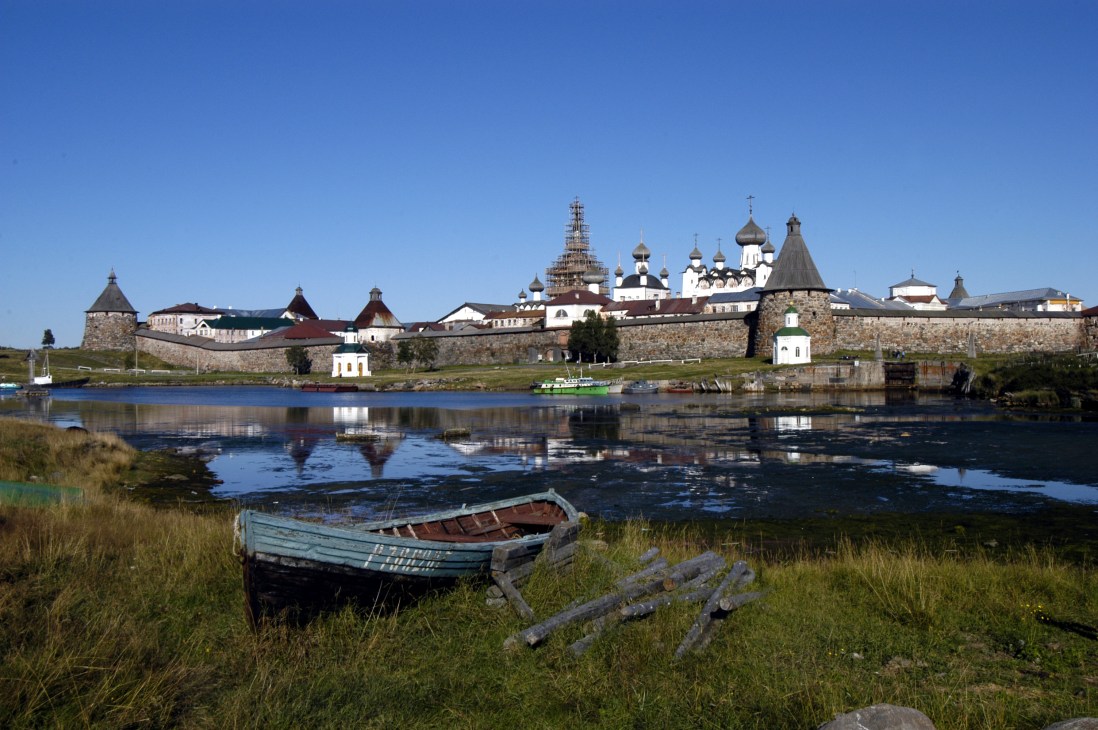 Solovki monastery with white and gold Orthodox domes reflected in calm waters of the White Sea, with a weathered blue rowboat in the foreground.