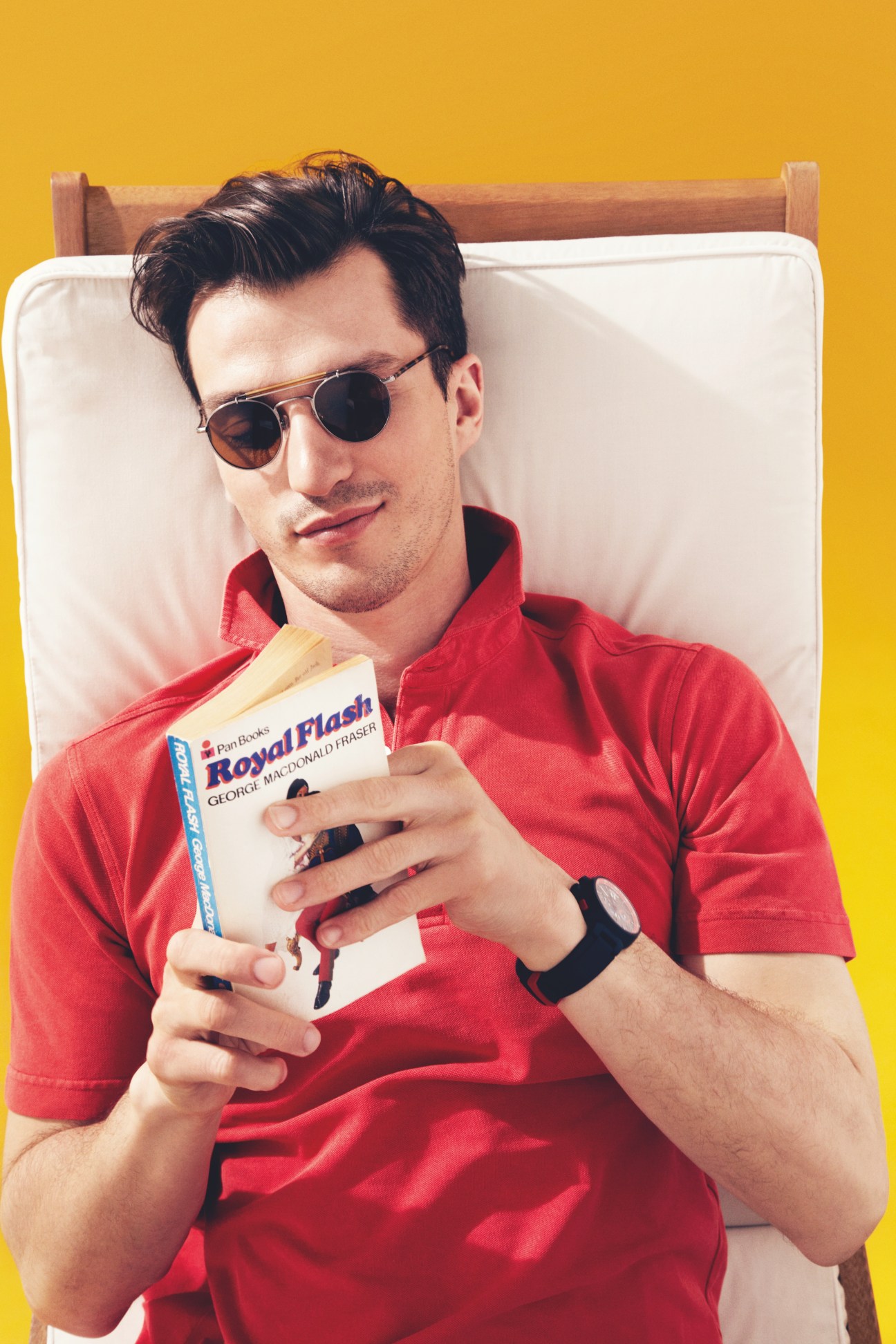 Man in red polo shirt wearing Oliver Peoples sunglasses and Louis Vuitton watch, lounging on bed with book against yellow background.