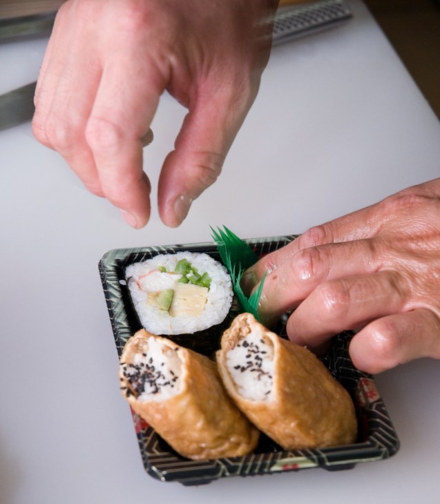 Hands holding a black tray displaying deep-fried tofu pouches and maki sushi with sesame seeds.