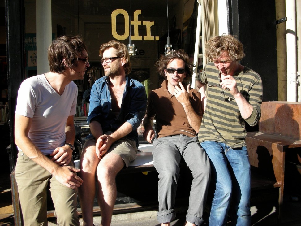Four men sitting outside the Ofr bookstore in Paris, conversing on a sunny day.