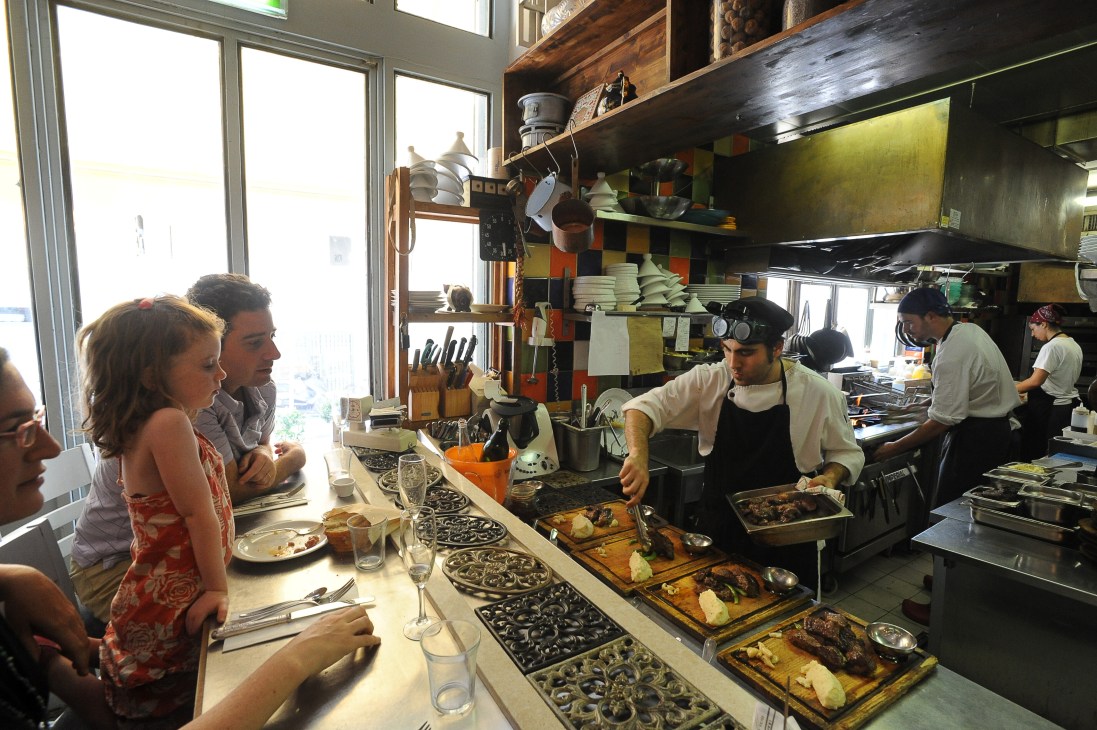 Interior of Machneyuda restaurant showing diners at counter with chefs working behind and open kitchen shelving.