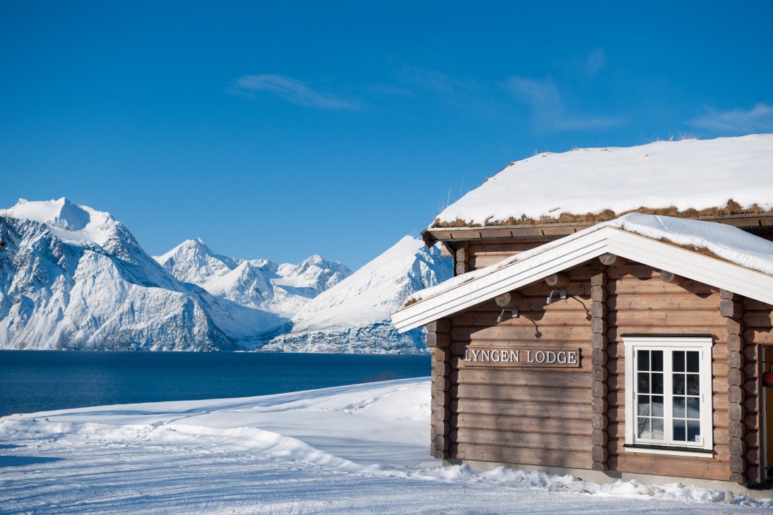 Lyngen Lodge wooden cabin overlooks snow-covered fjord with snow-capped mountains under blue sky.