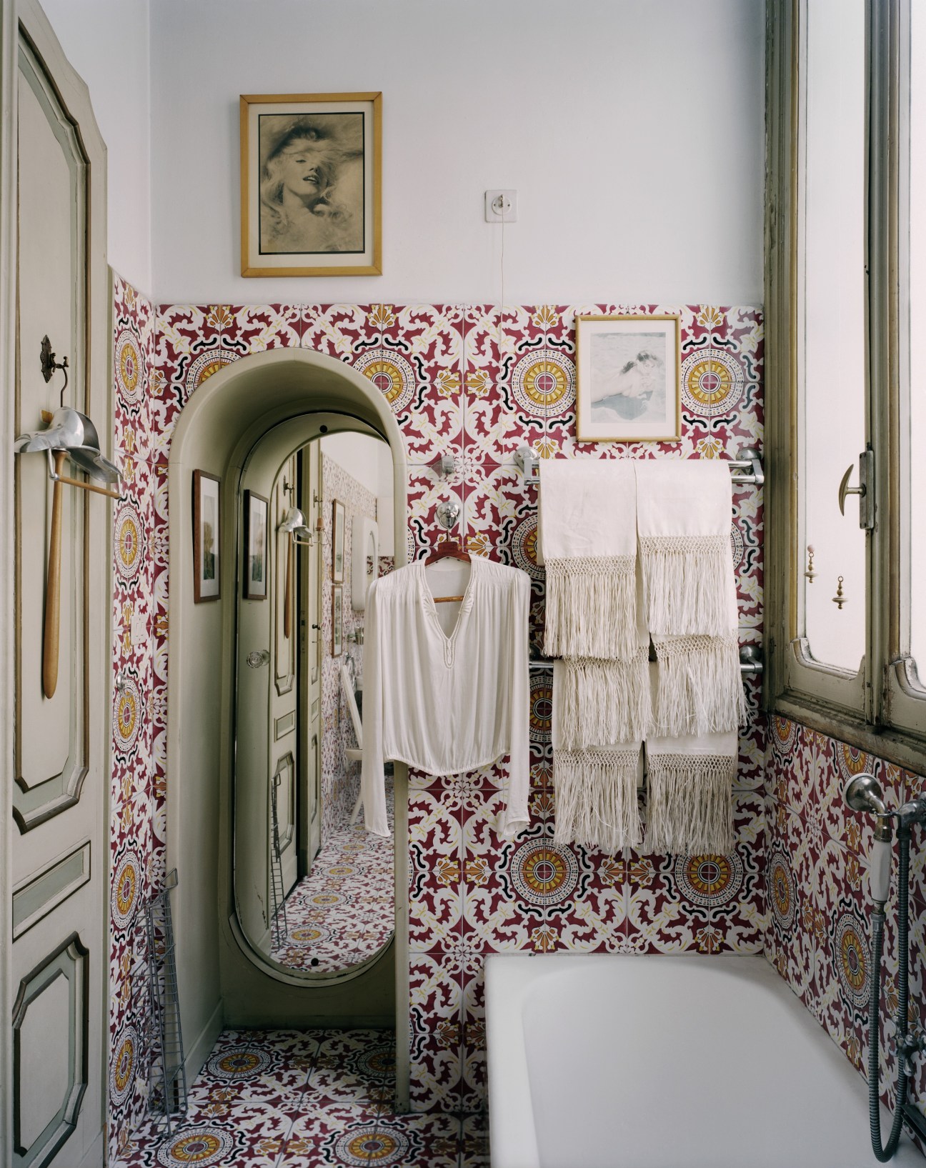 Ornate bathroom with red and gold patterned tiles, oval mirror, white towel rail and framed artwork.