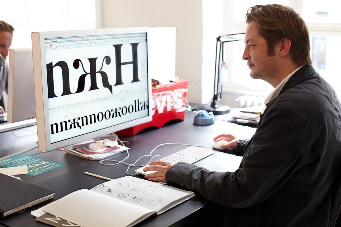 Man in dark jacket sits at desk viewing large computer monitor displaying typography design work.