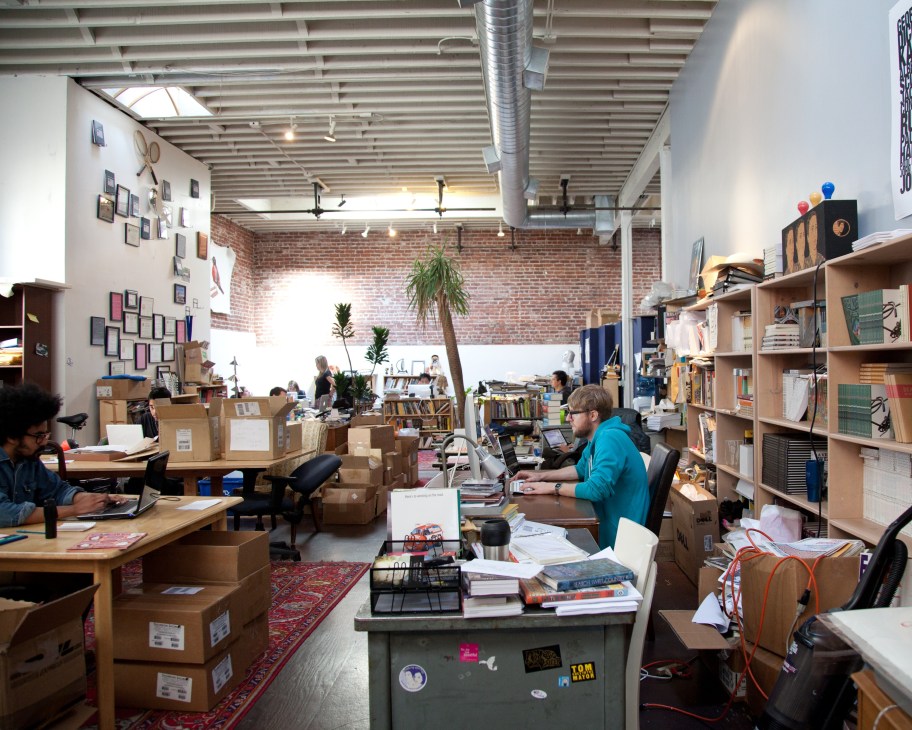Interior of The Believer and McSweeney's office with exposed brick, wooden shelves, cardboard boxes, and staff working at desks.