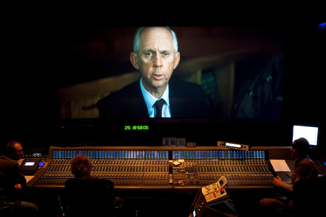 Dubbing engineers work at a large mixing console while monitoring a film scene on screen above.
