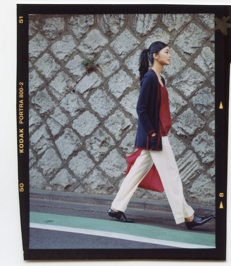 Woman in navy cardigan and white trousers walking past textured stone wall in Tokyo.