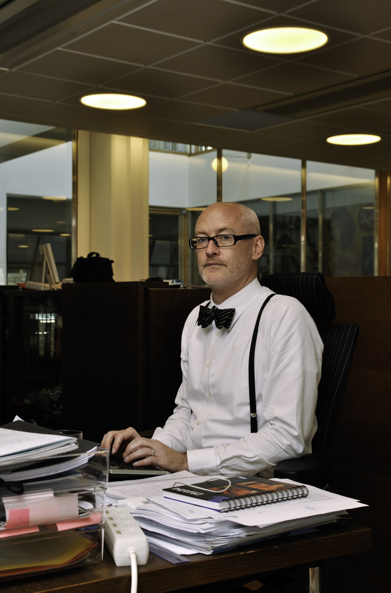 Man in white shirt and bow tie sitting at desk with papers and documents.