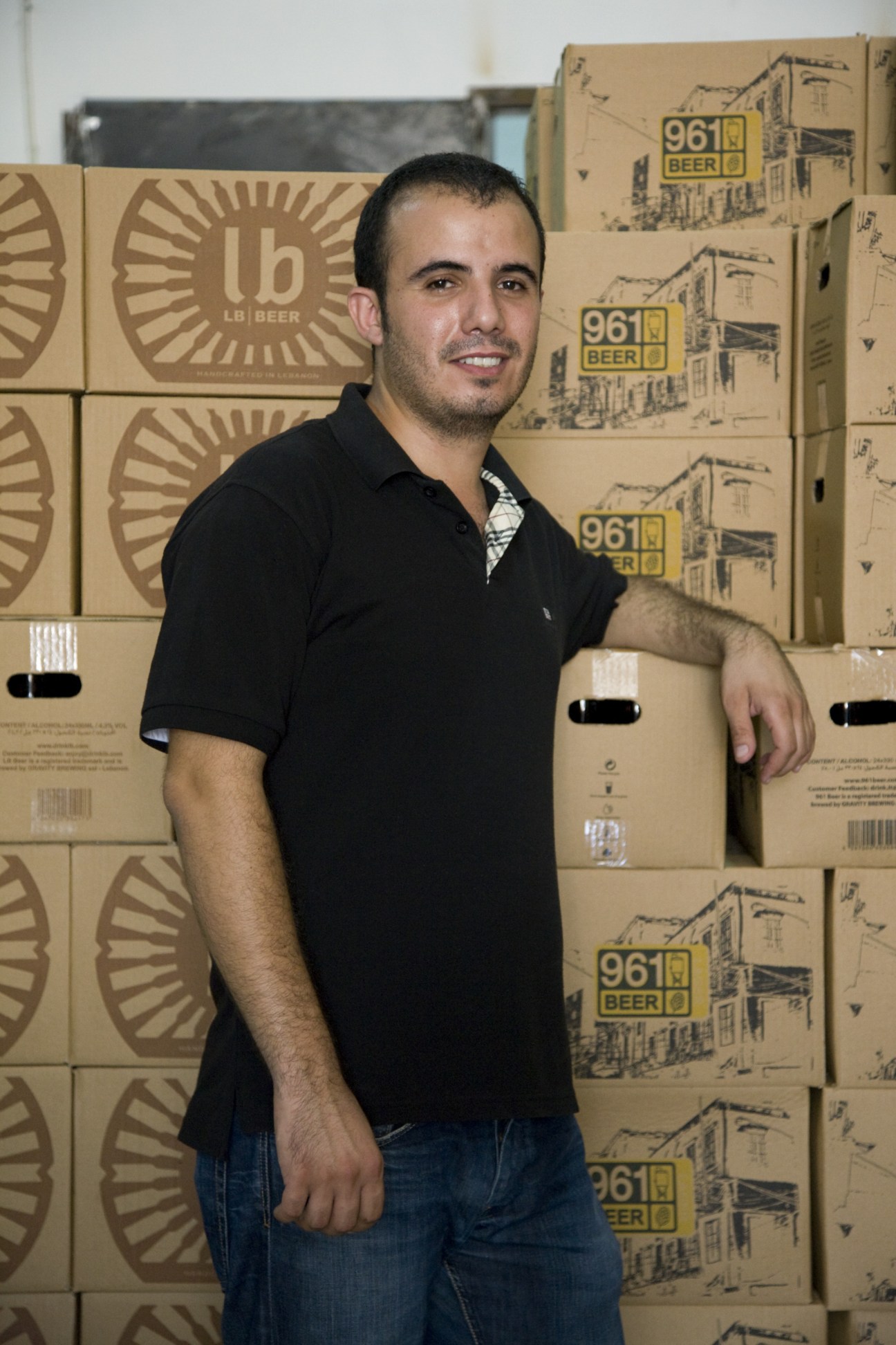 Man in black polo shirt posing with stacked cardboard boxes of LB and 961 beer.