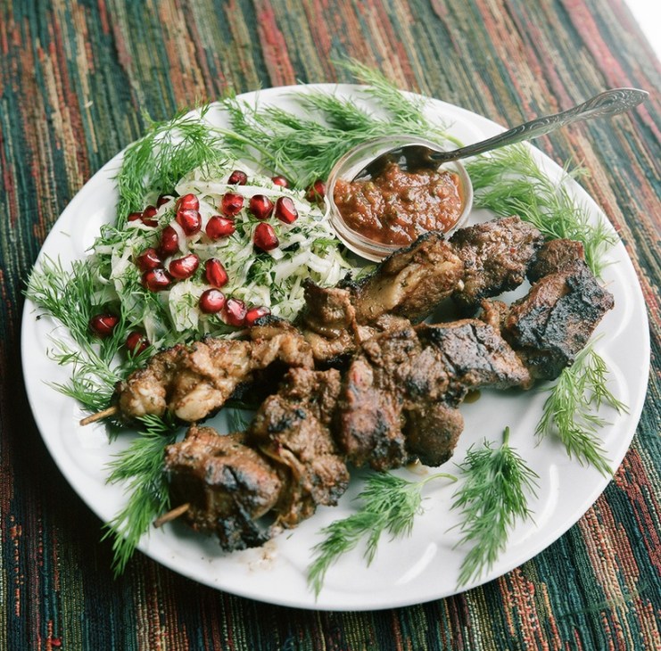 Grilled meat kebabs on skewers arranged on a white plate with dill, onions, pomegranate seeds and spicy sauce.