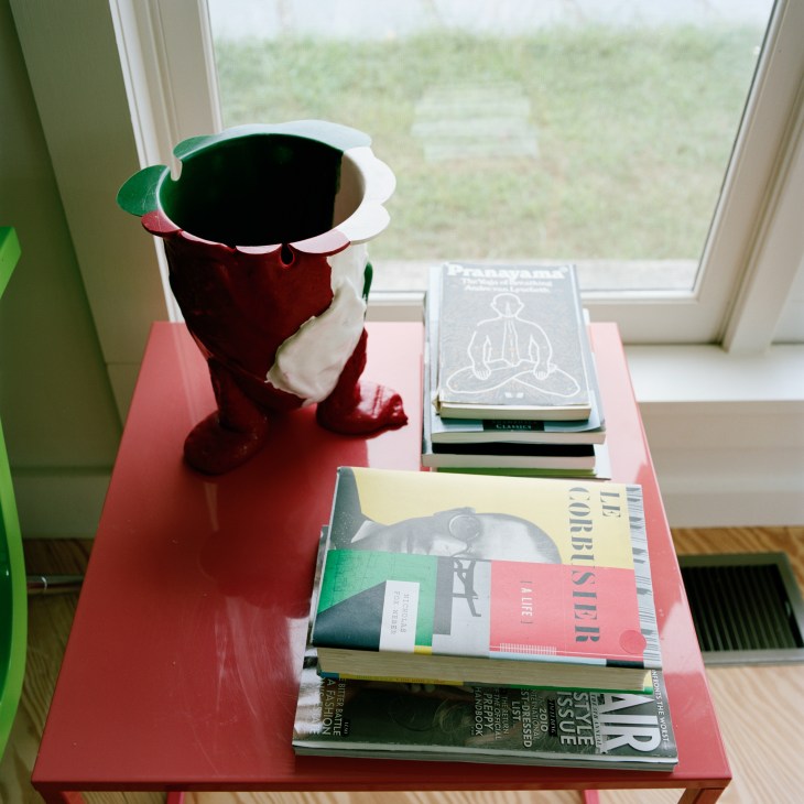 Books and magazines stacked on a pink table beside a decorative red and white vessel with a window view.