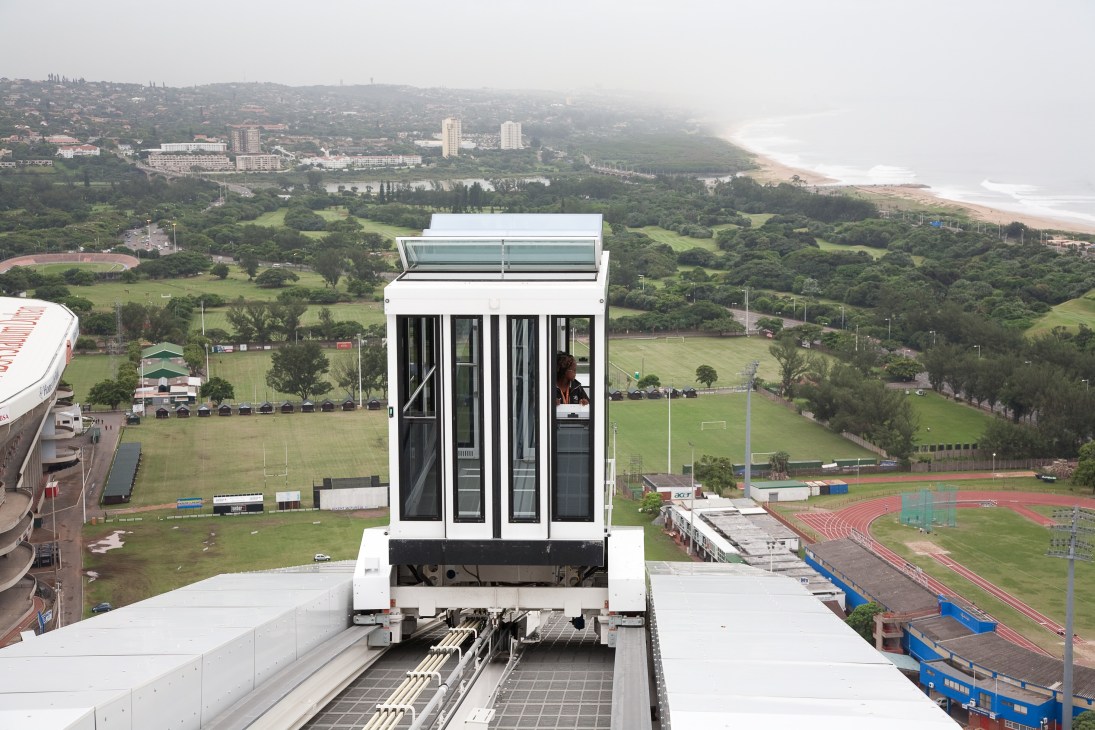Funicular cable car cabin suspended above Moses Mabhida stadium arch overlooking Durban coastal landscape.