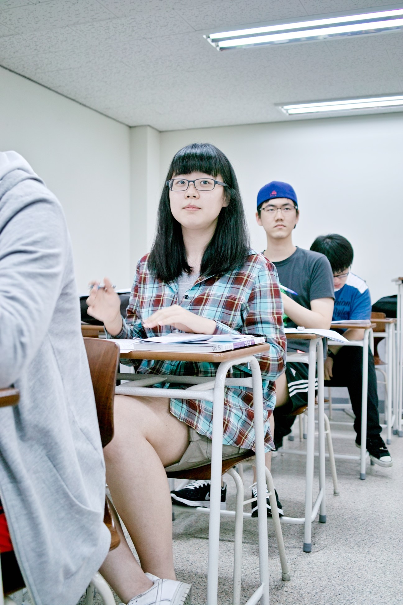 Students sit at desks in a fluorescent-lit classroom, with a girl in glasses and plaid shirt in the foreground.
