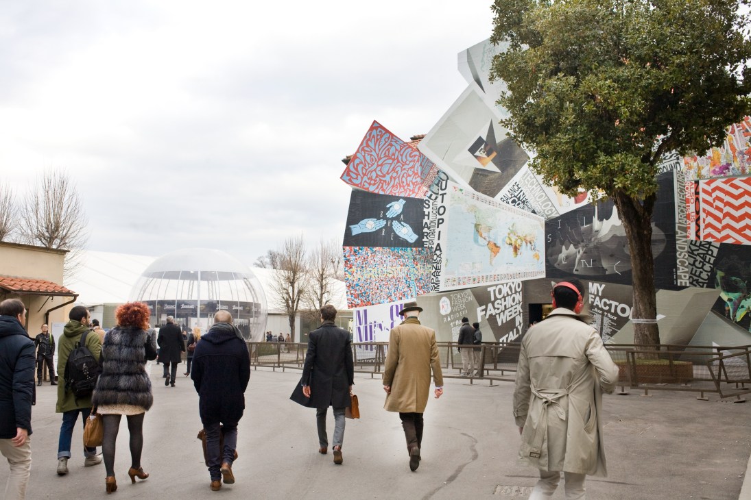 Visitors walk through Pitti Uomo exhibition grounds with colorful branded banners and signage visible.