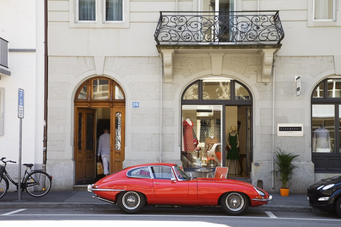 A red vintage sports car parked on a Zurich street lined with classical stone buildings and boutique shops.