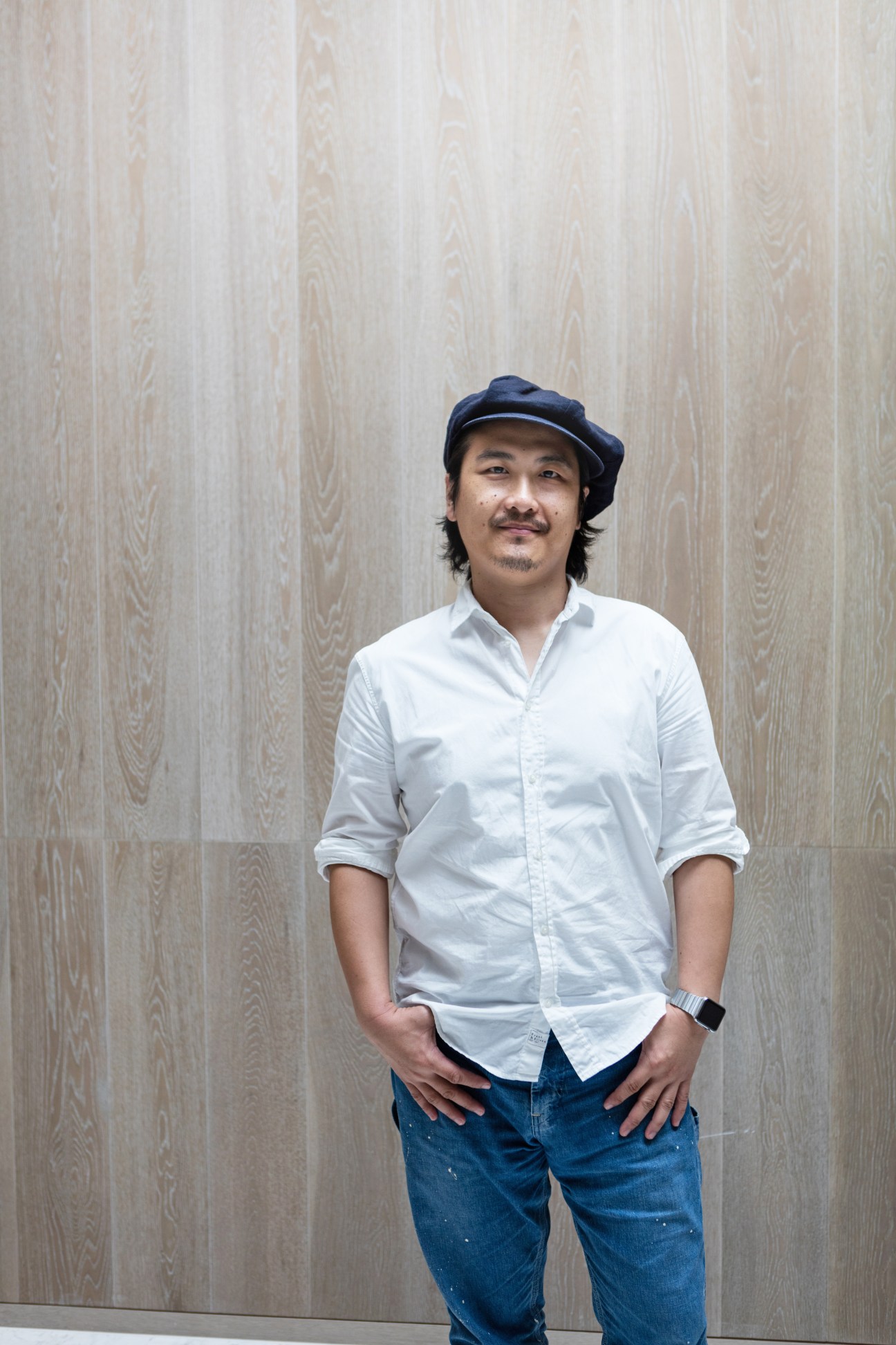 Man in white shirt and blue cap standing against wooden wall backdrop.