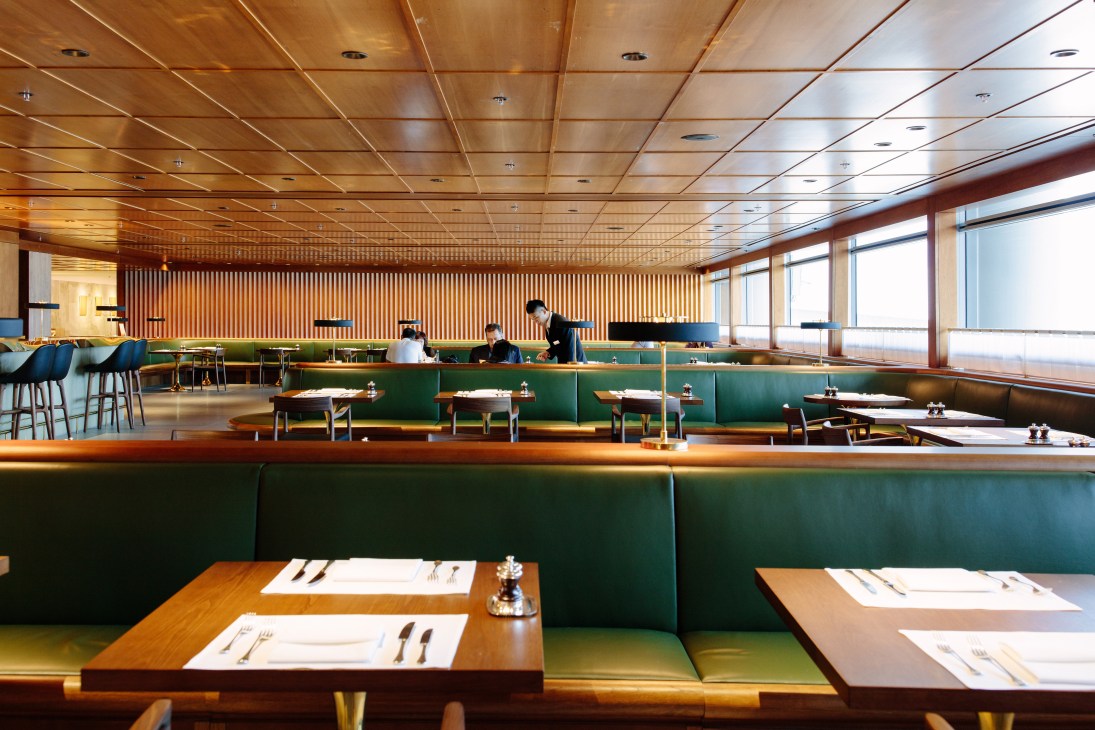 Modern restaurant interior with green leather banquette seating, wooden tables, and wood-paneled ceiling at Cathay Pacific first-class lounge.