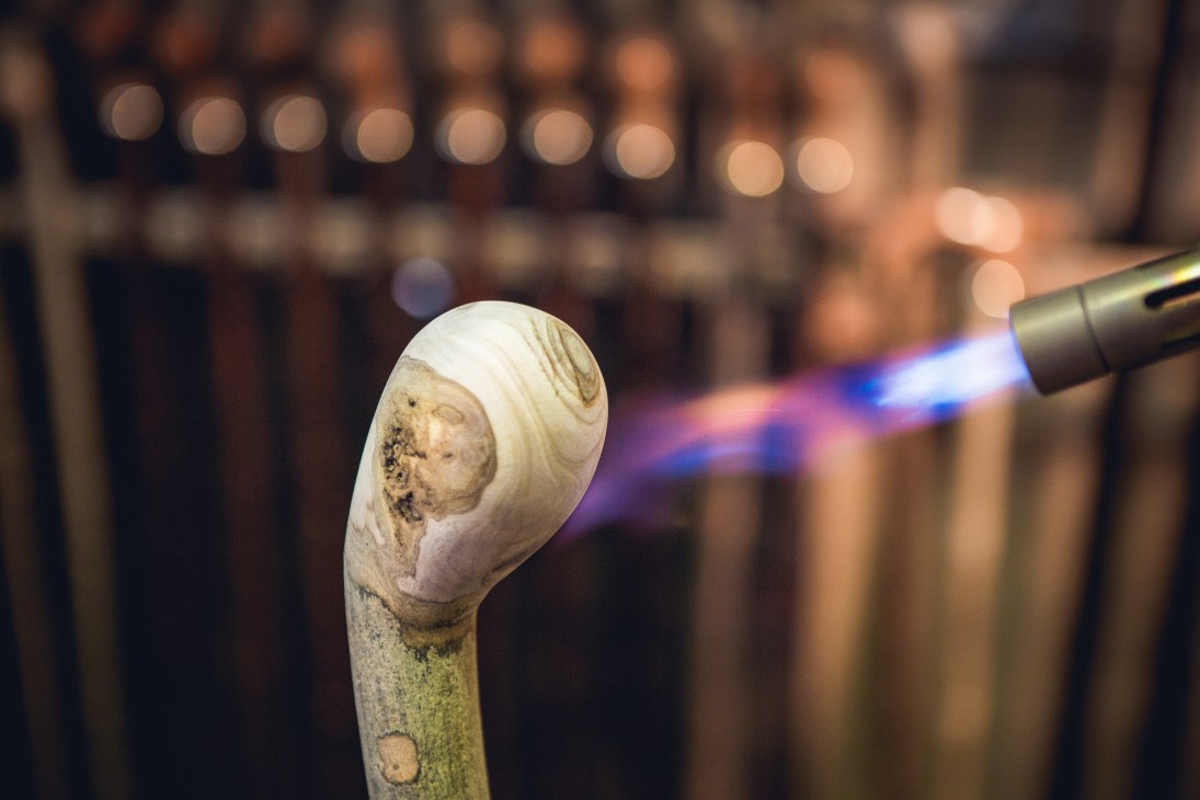 A wooden walking stick head being scorched with a blowtorch to accentuate its natural grain.
