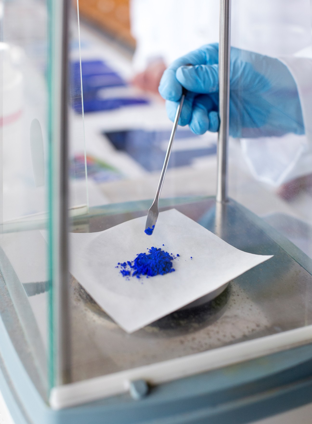 Researcher in blue gloves weighing blue pigment powder on precision scales in a laboratory fume hood.